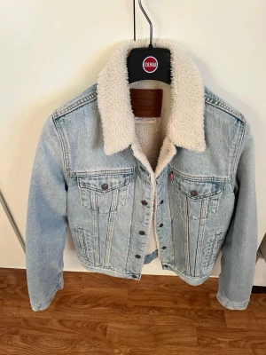 Levi's ljusblå sherpa jeansjacka - Säljer en ljusblå jeansjacka från Levi's med vit, fluffig sherpa-foder och krage. Klassisk modell med två bröstfickor, knappstängning och detaljerade sömmar. Perfekt för dig som vill ha en ikonisk och trendig jacka med extra värme.