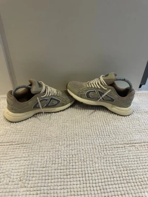 Beige Dior sneakers med mesh och läder - Snygga beige sneakers från Dior med vita och grå detaljer. Skorna har en chunky sula, meshpaneler för extra ventilation och läderdetaljer. Klassisk snörning och tydligt Dior-märke på plösen. Perfekta för dig som gillar exklusiva och trendiga sneakers.