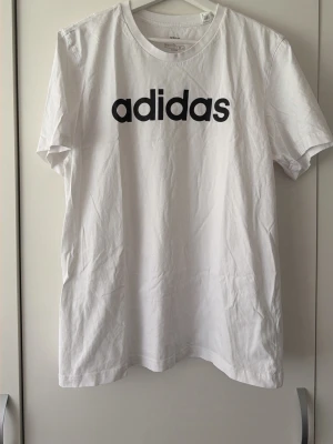 Vit adidas t-shirt med logga - Klassisk vit t-shirt från adidas med stor svart logga framtill. T-shirten har rund halsringning och korta ärmar. Tillverkad i mjuk bomull för en skön känsla och enkel stil.