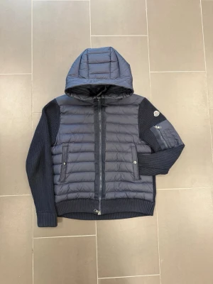Mörkblå pufferjacka från Moncler - Snygg mörkblå pufferjacka från Moncler med stickade ärmar och quiltad kropp. Jackan har huva, två fickor med knapp framtill och en Moncler-logga på ärmen. Perfekt för somriga dagar och riktigt trendig look.