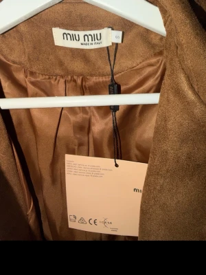 Brun trenchcoat från Miu Miu - Snygg brun trenchcoat från Miu Miu i mockaliknande material. Jackan har klassisk krage, knäppning framtill och ett matchande knytskärp i midjan. Perfekt för dig som vill ha en stilren och trendig look med exklusiv känsla.