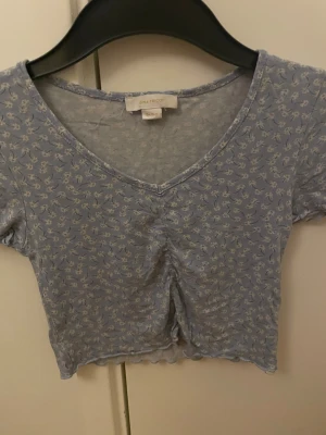 Blommig croppad topp från Gina Tricot - Söt ljusblå croppad topp från Gina Tricot med små vita blommor, v-ringning och korta ärmar. Tillverkad i mjuk bomull och har en lätt rynkad detalj framtill. Perfekt för dig som gillar en feminin och trendig stil.