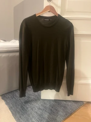 Olivgrön stickad tröja J.Lindeberg - Snygg olivgrön stickad tröja från J.Lindeberg med rund halsringning och långa ärmar. Tröjan är tunnstickad och har en klassisk, stilren design som passar perfekt till olika outfits. Tillverkad i 100% ull.  