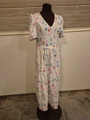 Blommig sommarklänning - Söt vit klänning med blommigt mönster🤍🌸🌼 Lätt och sval när det är varmt. Egentligen har den en avslappnad passform men om man vill ha den tajt i midjan är den enkel att knyta i midjan med ett band eller fästa med en broch. Står S men passar M och kanske L också. Använd en gång så är i princip nyskick. Det är bara att skriva om man har frågor!😊💖