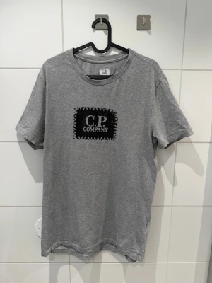 C.P. Company - Stitch Logo Patch T-shirt - Snygg grå t-shirt från C.P. Company med ett svart tryck och logga på bröstet. Klassisk rund hals och korta ärmar. Tillverkad i mjuk bomull som känns skön mot huden. Perfekt för en avslappnad och trendig look. Passar mig som har storlek L