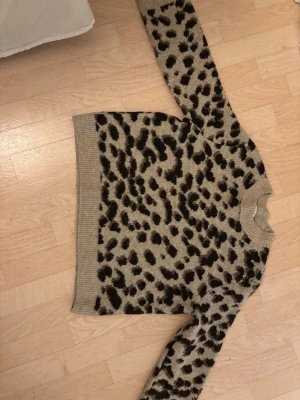 Beige leopardmönstrad stickad tröja H&M - Stickad tröja från H&M i beige med svart och brunt leopardmönster. Tröjan har ribbstickad krage, ärmslut och nederkant. Perfekt för dig som gillar djurmönster och vill ha en mysig och trendig look.