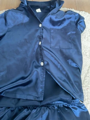 Mörkblå satinpyjamas med kort ärm - Snygg mörkblå pyjamas i glansigt satinmaterial. Setet består av en kortärmad skjorta med vita knappar och bröstficka samt matchande shorts med resår i midjan. Perfekt för mysiga kvällar hemma.