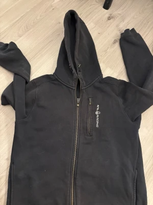 Svart hoodie från Sail Racing - Svart hoodie från Sail Racing med dragkedja framtill och bröstficka med logga. Utrustad med huva och justerbara snören. Insidan är mjuk och fleecefodrad för extra komfort. Perfekt för dig som gillar en sportig och stilren look.