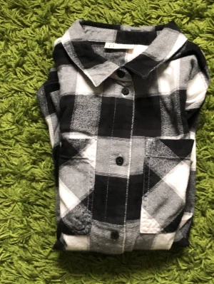 Svartvit rutig skjorta med knappar - Klassisk svartvit rutig skjorta med lång ärm och knappar framtill. Skjortan har två bröstfickor och är tillverkad i mjuk bomull, perfekt för en avslappnad och stilren look.