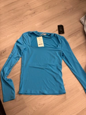 Blå långärmad topp från NA-KD - Snygg blå långärmad topp från NA-KD med fyrkantig ringning. Tillverkad i mjukt och stretchigt material som sitter skönt på kroppen. Perfekt att styla med jeans eller kjol för en trendig look.