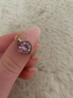 Guldfärgad ring med rosa sten - Snygg guldfärgad ring med en stor, rund rosa sten i mitten. Ringen har en enkel men elegant design som verkligen låter stenen stå i fokus. Perfekt för dig som vill ha en färgklick på handen och gillar unika accessoarer.