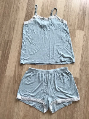 Turkost pyjamas-set med spetsdetaljer från KappAhl, stl 146/152 - Sött turkost pyjamas-set med linne och shorts, stl 146/152. Linnet har tunna axelband och vit spets längs ringningen. Shortsen har elastisk midja och matchande vit spets vid bensluten. Tunnt, svalt ribbat tyg. Perfekt för mysiga kvällar hemma. På bilden väldigt skrynkliga, men i fint skick. Just nu fri frakt, så du betalar bara för pyjamasen. Jag har fler plagg ute, och hittar du något mer från min garderobsutrensning, så skickar jag allt i samma paket.