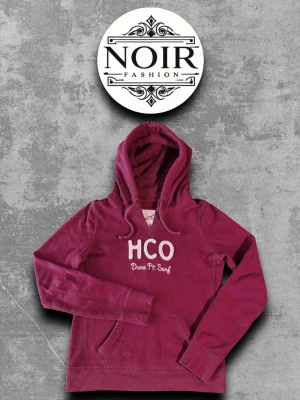 Vinröd hoodie från HCO - Snygg vinröd hoodie från Hollister med broderad HCO-logga och texten Dana Pt. Surf på bröstet. Hoodien har en klassisk känguruficka framtill och justerbar huva med dragsko. Perfekt för en avslappnad och trendig look.