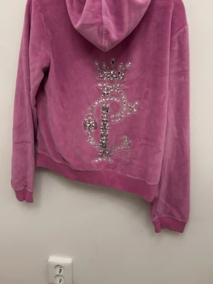 Rosa velour set  från Pierre Cardin i strl 42 - Mjuk rosa hoodie i velour från Pierre Cardin med dragkedja framtill och två fickor. Baksidan har ett stort glittrigt emblem med krona och bokstäver i strass. Perfekt för en avslappnad och trendig look.