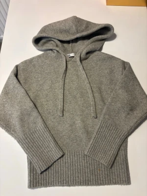 Grå stickad hoodie med ribbade detaljer - Mysig grå stickad hoodie med ribbade muddar och nederkant. Tröjan har en avslappnad passform och en stor huva som ger en cool look. Perfekt för att hålla sig varm under kyliga dagar❄️❄️