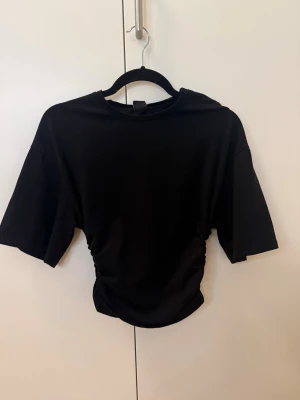 Svart croppad topp med rynkade sidor - Hej! Säljer min snygga svarta croppad topp med rynkade detaljer på sidorna och korta ärmar. Tillverkad i mjuk bomull som känns skön mot huden. Perfekt till jeans eller kjol för en trendig look. Skriv om det är något ni undrar!🩷