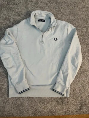 Ljusblå halvzip från Fred Perry - Snygg ljusblå stickad tröja från Fred Perry med halv dragkedja och broderad logga på bröstet. Tröjan har ribbade muddar och krage, samt diskreta mörkblå detaljer vid ärmslut och nederkant. Perfekt för lager på lager och stilren look.