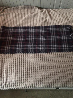 Mörkblå rutig halsduk från Burberrys - Säljer en klassisk mörkblå halsduk från Burberrys med rutigt mönster i vitt och rött. Halsduken är tillverkad i 100% lammull och har fransar i ändarna. Perfekt för dig som vill ha en tidlös accessoar med brittisk känsla. Den är helt ny och är i perfekt skick!