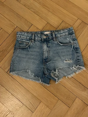 Blå jeansshorts från Gina Tricot - Säljer ett par blå jeansshorts från Gina Tricot med fransiga kanter och slitna detaljer. Klassisk femficksmodell med knapp och dragkedja framtill. Perfekta till sommaren och ger en avslappnad look.