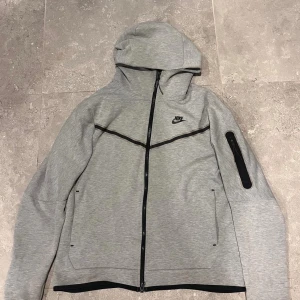 Grå nike tech - Grå vindjacka från Nike med svart dragkedja och huva. Jackan har en stilren design med svarta detaljer, inklusive en ficka med dragkedja på ärmen. Perfekt för dig som gillar sportig och clean look.