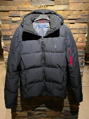 Svart pufferjacka från Polo Ralph Lauren unik s äkta  - Då jag har för många jackor måste några bort. Är i nyckick och köptes från Ralph lauren för ca 4100 kr. Kvitto finns som går att verifiera!! Storlek S men passar även M! Pris går att diskutera men skriv inte ifall du ej har pengarna. Snygg svart pufferjacka från Polo Ralph Lauren med huva och dragkedja framtill. Jackan har en röd detalj på ärmfickan och diskret logga på bröstet. Perfekt för kalla vinterdagar och riktigt skön att bära.