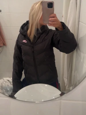 Svart pufferjacka från Nike - Säljer en svart pufferjacka från Nike med huva och dragkedja framtill. Jackan har en rosa Nike-logga på bröstet och är vadderad för extra värme. Perfekt för kalla dagar och har en modern, oversized passform.