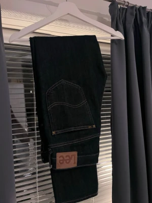 Mörkblå Lee Daren jeans W31 L32 - Säljer ett par mörkblå Lee Daren jeans med klassisk femficksdesign och brunt Lee-märke på bakfickan. Jeansen har dragkedjegylf och är tillverkade i bomull med en snygg, tidlös passform. Perfekta för dig som gillar stilrena och bekväma jeans. Använda enbart någon vecka bara. Lätt att styla med!