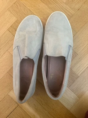 Beige slip-on sneakers  - Snygga slip-on sneakers från DASIA i grå/beige mockaimitation med vit sula. Skorna har elastiska paneler på sidorna för enkel påtagning och en stilren, minimalistisk design. Perfekta för en avslappnad och trendig look.