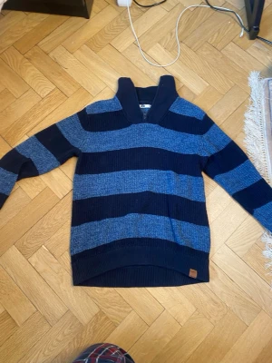 Blå randig stickad tröja från WE - Säljer en stickad tröja från WE med breda horisontella ränder i mörkblått och ljusblått. Tröjan har en hög krage med dragkedja och ribbstickade muddar vid ärmslut och nederkant. Perfekt för att hålla sig varm under kyliga dagar.
