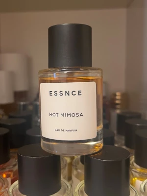 ESSNCE Hot Mimosa Eau de Parfum - ESSNCE Hot Mimosa Eau de Parfum i en stilren, rund glasflaska med svart lock. Parfymen har en varm, gyllene färg och minimalistisk etikett. Perfekt för dig som gillar moderna och fräscha dofter.