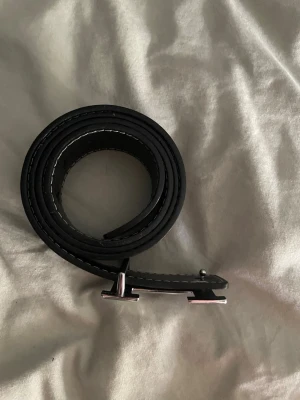 Svart skärp med silverfärgat H-spänne - Snyggt svart bälte med ett stilrent silverfärgat H-format spänne. Bältet har vita sömmar längs kanterna och är gjort i slätt skinn. Perfekt accessoar för att lyfta din outfit.