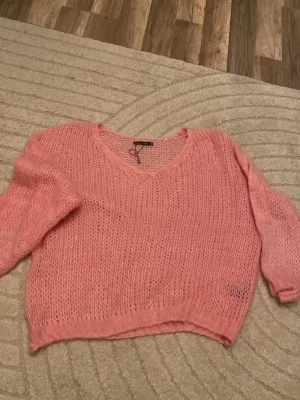 Rosa stickad tröja från Black Colour - Säljer en oversized stickad tröja i en härlig rosa färg från Black Colour. Tröjan har v-ringning, långa ärmar och är stickad i ett luftigt, grovt garn som ger en avslappnad look. Perfekt att slänga på sig när det är kyligt ute.