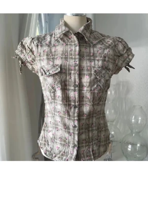 Rutig kortärmad skjorta med rosor - Kortärmad skjorta i beige och brunt rutigt tyg med små rosa rosor. Skjortan har knappar framtill, två bröstfickor med lock och knappar samt puffärmar med knytband. Figurnära passform och klassisk krage. Perfekt för en söt och avslappnad stil.