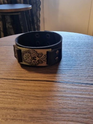 Armband vintage viking - Svart läderarmband med ett detaljerat vikingmotiv i metall. Armbandet har justerbara tryckknappar för att passa olika handledsstorlekar. Storlek 23 cm långt. Bredd 3 cm.