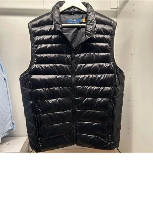 Svart dunväst från Polo Ralph Lauren - Svart dunväst från Polo Ralph Lauren med klassisk quiltad design och dragkedja framtill. Västen har två fickor med dragkedja och ikonisk broderad logga på bröstet. Perfekt för lager-på-lager och ger en stilren look.