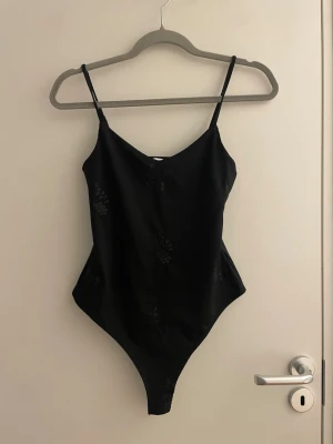 Svart body med tunna axelband - Snygg svart body med tunna axelband och diskreta blommiga spetsdetaljer. Kroppsnära passform och v-ringad framtill. Perfekt att styla med jeans eller kjol för en trendig look.