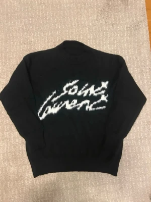 Svart Saint Laurent stickad tröja - Snygg svart stickad tröja från Saint Laurent med vit, fluffig logga i graffiti-stil på bröstet. Tröjan har rund hals och långa ärmar. Perfekt för dig som vill ha en trendig och exklusiv look. Materialet ser ut att vara mjukt och lite luddigt.