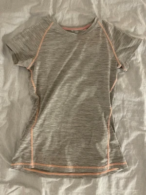 Grå sport-tshirt - Snygg och funktionell grå sport-tshirt med diskreta orange sömmar. T-shirten har rund halsringning och korta ärmar, tillverkad i ett mjukt och stretchigt funktionsmaterial som andas. Perfekt för träning eller gympass.❤️