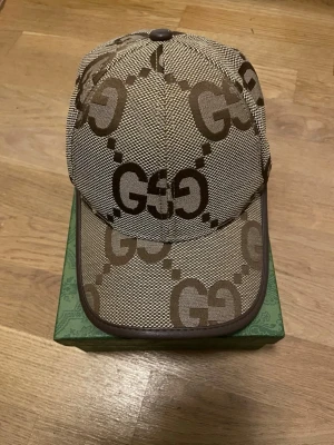 Beige GG keps från Gucci - Säljer en beige keps från Gucci med det klassiska GG-mönstret i brunt över hela kepsen. Kepsen har böjd skärm, justerbart band bak och detaljer i brunt läder. Perfekt för dig som vill ha en stilren och lyxig accessoar.