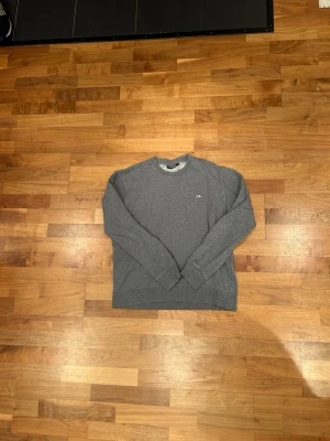 Grå sweatshirt från J.Lindeberg - En stilren grå sweatshirt från J.Lindeberg med rund halsringning och diskret logga på bröstet. Tröjan har långa ärmar och ribbade muddar vid ärmslut och nederkant. Perfekt för en avslappnad och snygg look.