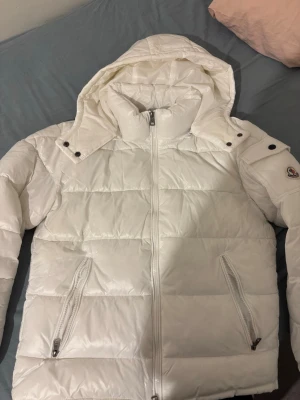 Vit dunjacka från Moncler med huva - Säljer en snygg vit dunjacka från Moncler med avtagbar huva och två dragkedjeförsedda fickor. Jackan har quiltad design och Moncler-logga på ärmen. Perfekt för kalla vinterdagar och tillverkad i glansigt material.