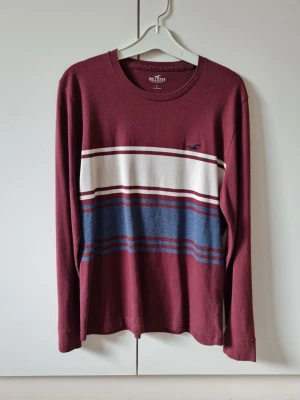 Vinröd långärmad tröja Hollister strl S - Snygg långärmad tröja från Hollister i vinrött med breda horisontella ränder i vitt och blått över bröstet. Klassisk crewneck och liten Hollister-logga broderad på bröstet. Material 100% bomull. Storlek S.