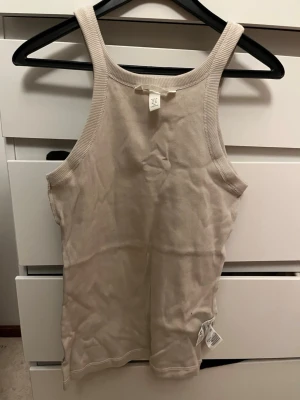 Beige ribbad linnetopp från H&M - Säljer en enkel och stilren beige linnetopp från H&M. Toppen är ribbad och har breda axelband samt rak halsringning. Perfekt att matcha med jeans eller kjol för en clean look.