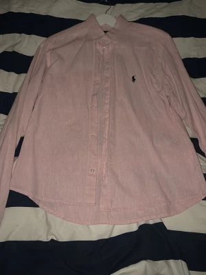 Ljusrosa skjorta från Ralph Lauren - Klassisk ljusrosa skjorta från Ralph Lauren med button-down krage och broderad logga på bröstet. Skjortan har lång ärm och knäppning framtill. Tillverkad i mjuk bomull för en bekväm känsla och stilren look.