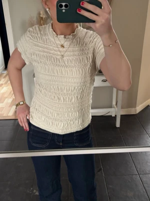 Krämvit ribbad topp med kort ärm - Säljer en krämvit ribbad topp med korta ärmar och rund hals. Toppen har ett smockat mönster som ger en snygg struktur och sitter figurnära. Perfekt att styla med jeans eller kjol för en trendig look. Från Hm! Storlek M men sitter tajtare så skulle säga S. 