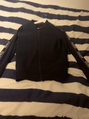 Svart hoodie från Hugo med dragkedja - Snygg svart hoodie från Hugo boss med hel dragkedja framtill och logga på ärmarna. Klassisk passform och ribbade muddar. Perfekt för en avslappnad och trendig stil. Materialet känns mjukt och bekvämt, troligen bomull eller en bomullsblandning. Och såklart äkta, var inte blyga med att kontakta eller ställa frågor om ni har några funderingar!