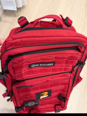 Röd ryggsäck från GASP - Cool röd ryggsäck från GASP med flera fack och svarta detaljer. Utrustad med patchar som 'NO EXCUSES', 'IRON WORLD' och 'BETTER BODIES'. Tillverkad i slitstarkt material och har justerbara remmar samt spännen för extra funktionalitet.