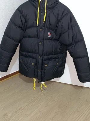 Svart dunjacka från Fjällräven - Snygg svart dunjacka från Fjällräven med gula dragskor och tryckknappar. Jackan har huva, två stora fickor framtill och klassisk Fjällräven-logga på bröstet. Perfekt för kalla dagar och riktigt varm tack vare dunfyllningen.