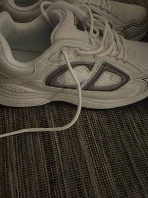 Vita Dior sneakers med grå detaljer - Snygga vita sneakers från Dior med gråa detaljer och tydlig CD-logga på sidan. Skorna har en chunky sula och är tillverkade i mesh och syntetmaterial för en modern look. Perfekta för dig som vill soppa upp din streetstyle med exklusiv känsla.