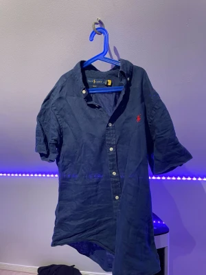 Blå linneskjorta från Ralph Lauren - Snygg mörkblå skjorta från Ralph Lauren i slim fit-modell. Tillverkad i linne med klassisk krage, korta ärmar och knappar framtill. Ikoniska röda loggan broderad på bröstet. Perfekt för en stilren och avslappnad look. 